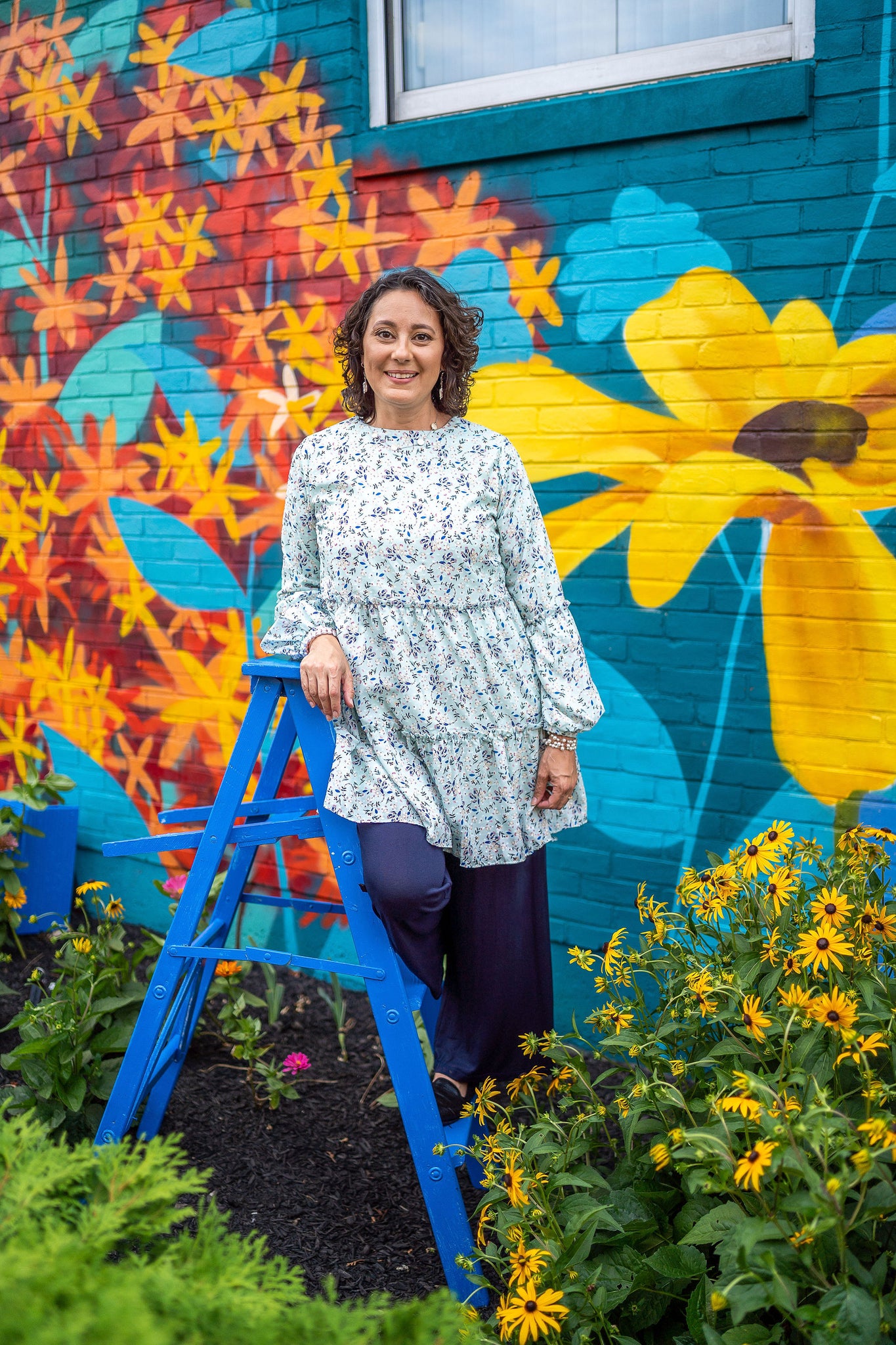 Woman leaning on ladder wearing navy blue pants and teal floral print long sleeve top